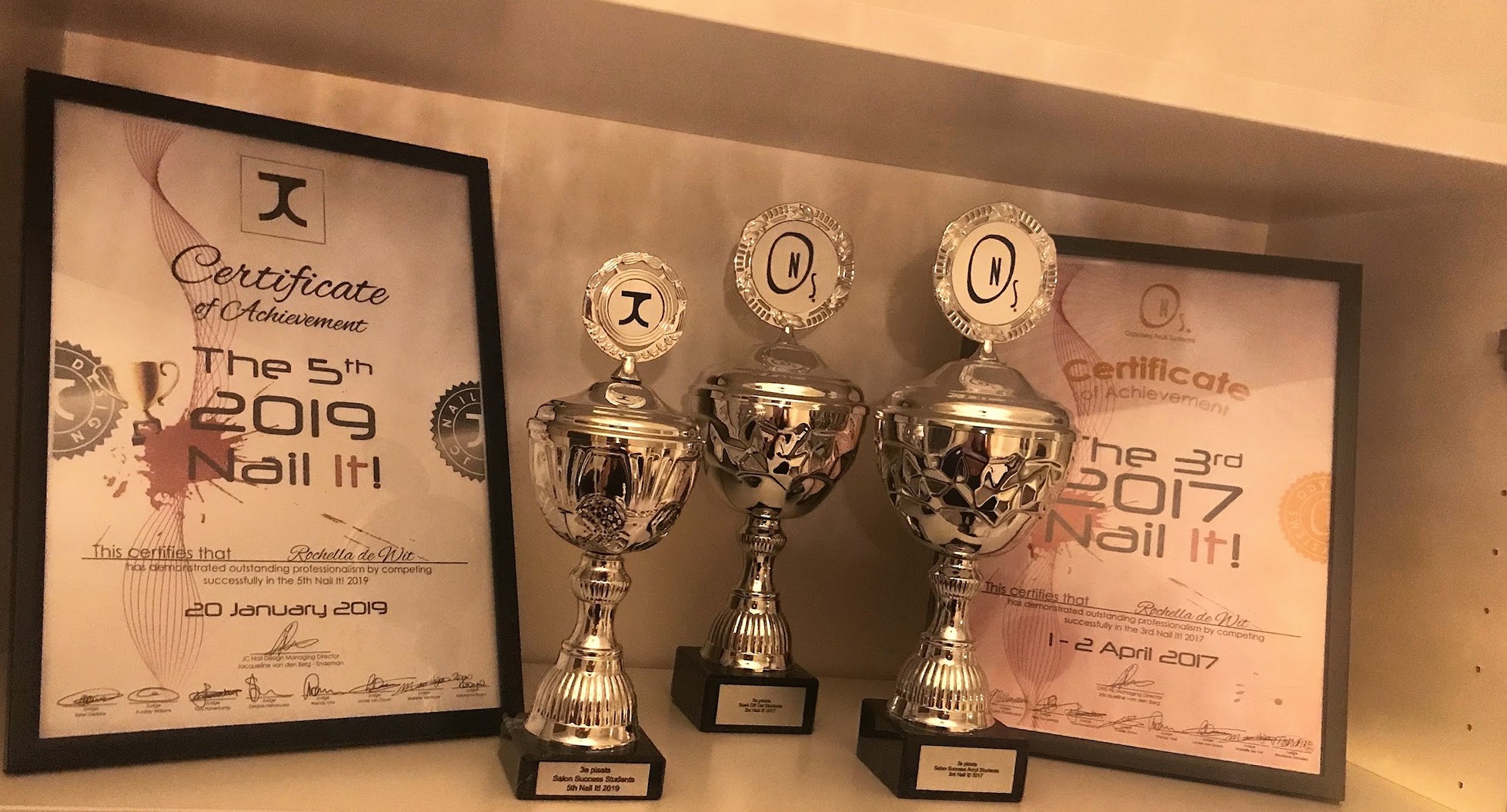 Twee certificaten van de Nail It!-wedstrijden van 2017 en 2019 met drie bekers ervoor